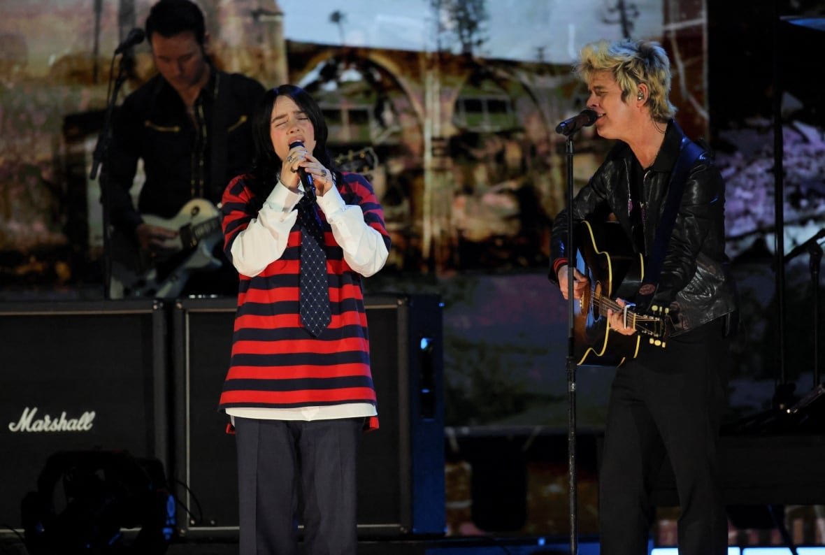 A woman wearing a black and red striped top with a tie sings on a stage with a man playing a guitar.