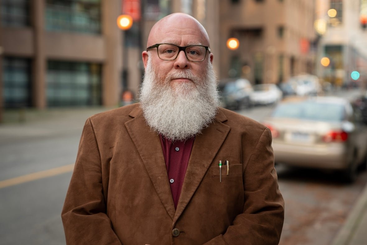 daniel-stern-chief-editor-driving-vision-news A bald man stands in the road in a brown jacket with a full, white beard and glasses.