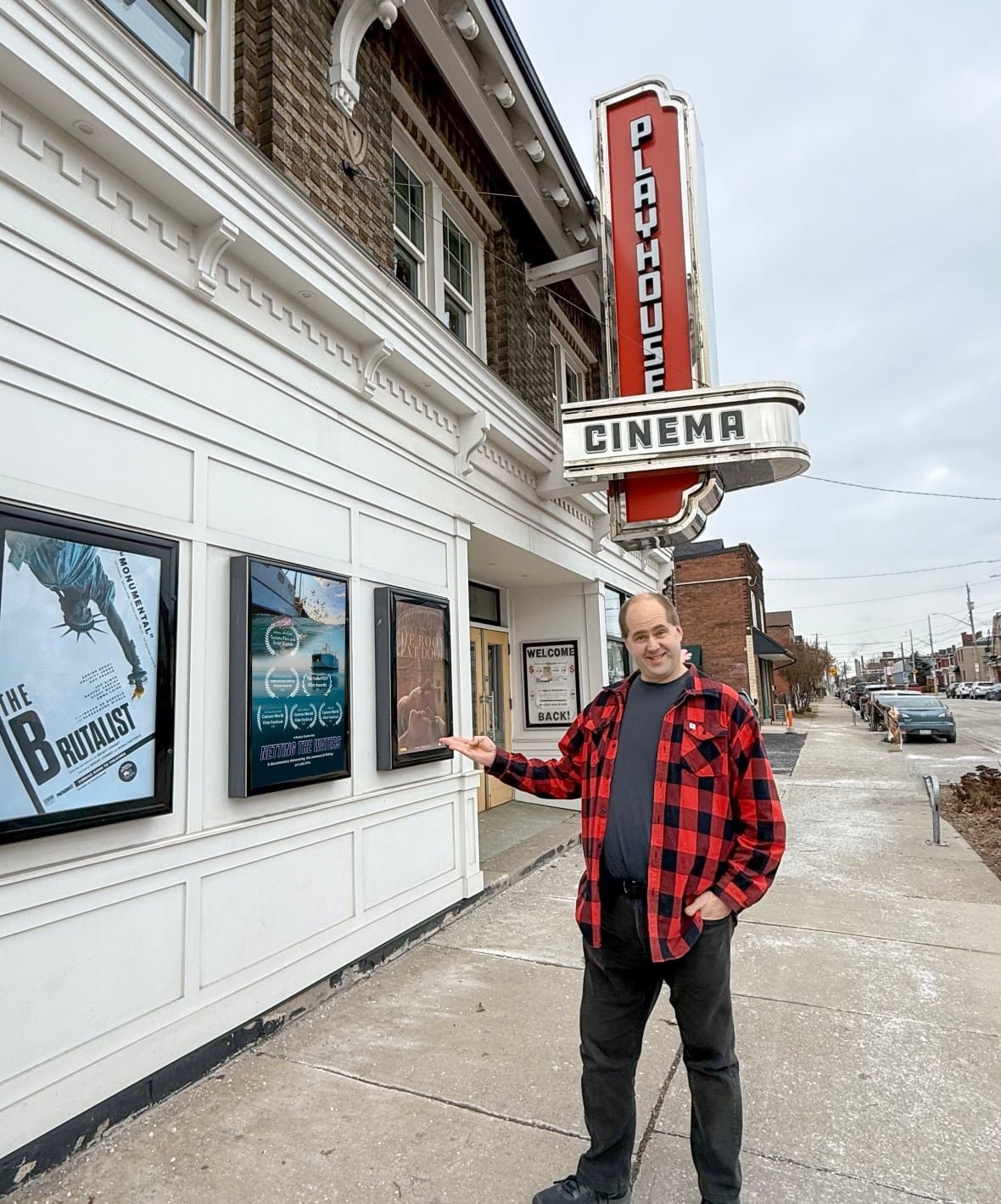 A person stands on a sidewalk by a theatre with a sign reading "Playhouse Cinema" and points toward the building.