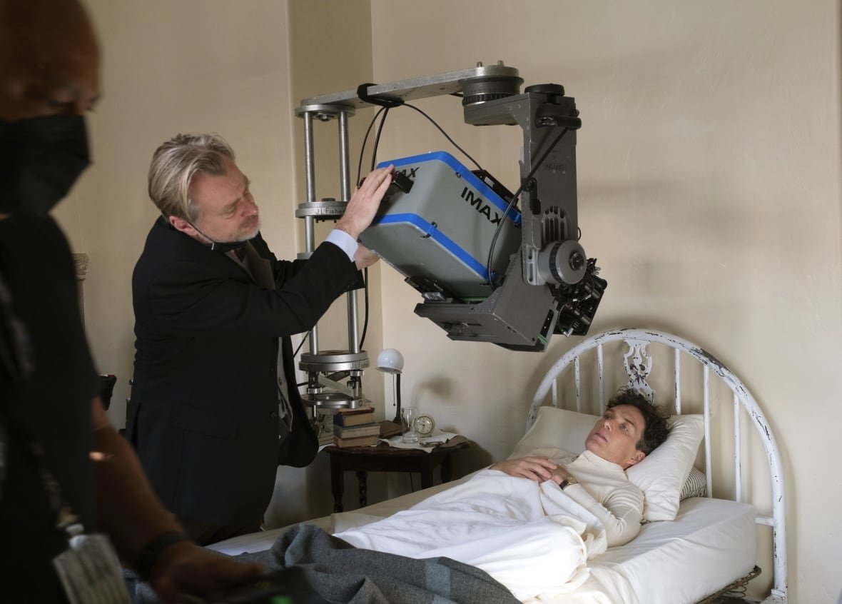 film-oppenheimer A blond man in a dark suit jacket adjusts an Imax camera hanging over a thin man in a bed.