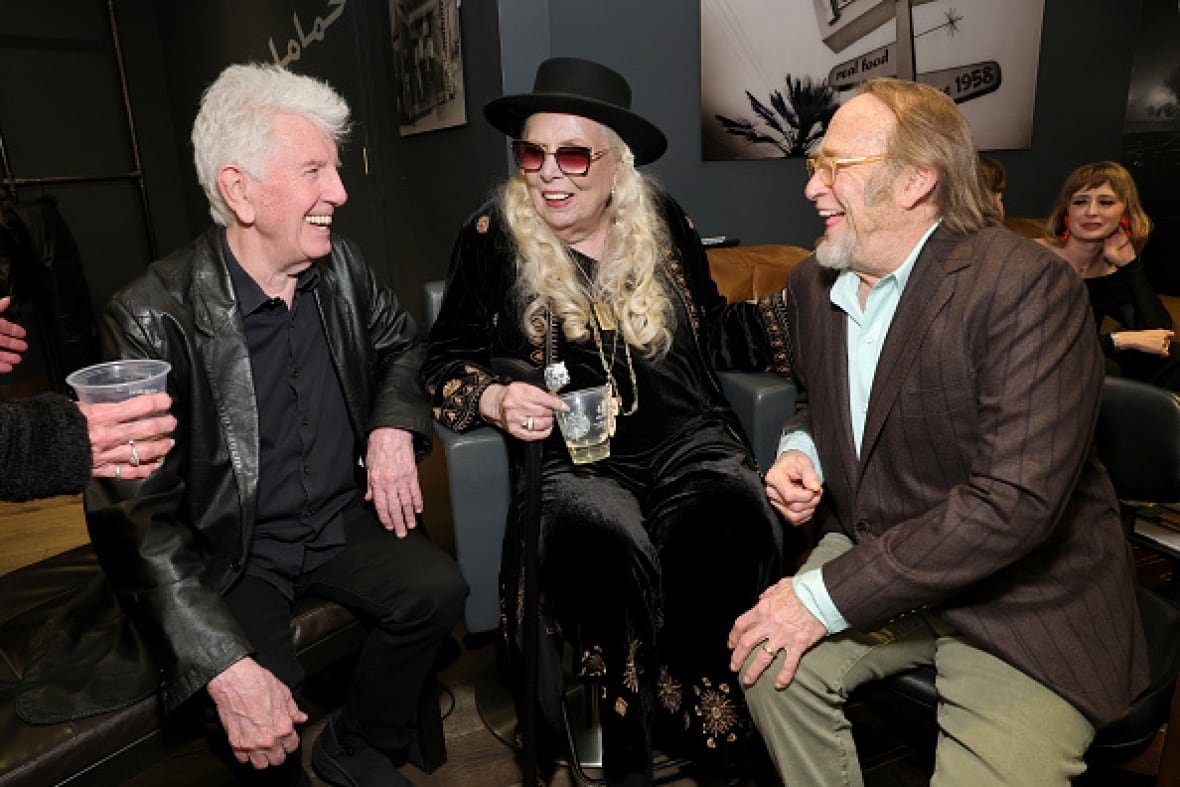 A woman with long blond hair sits on a chair. Two men sit on either side of her