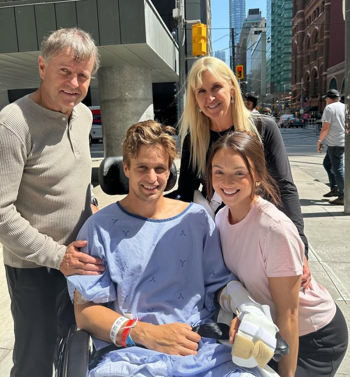 lucas-arsenault-and-family A man in a hospital gown and wheelchair, smiling with family.