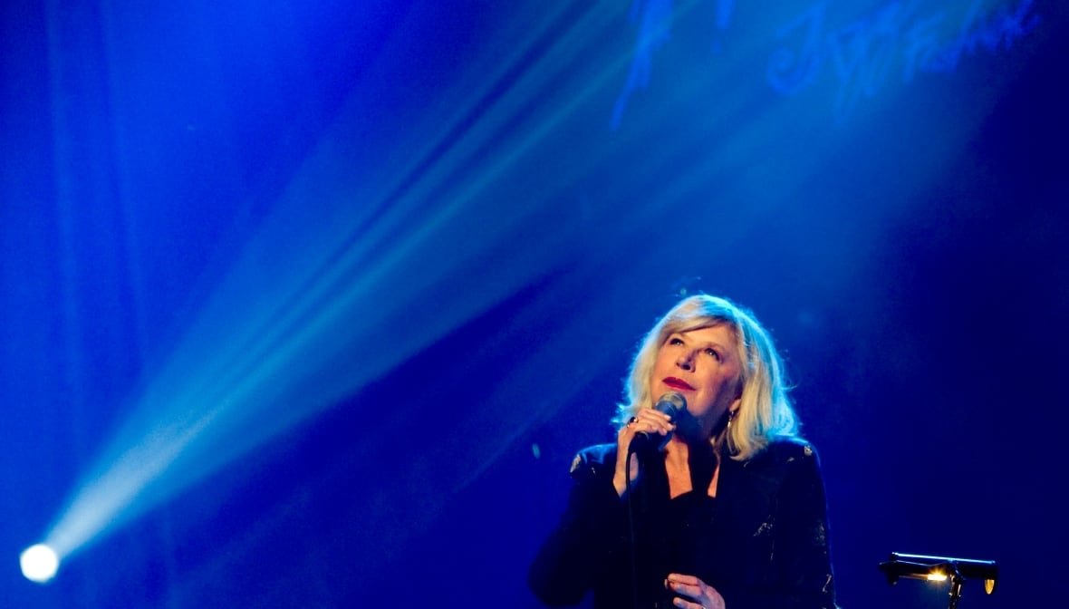 marianne-faithfull-obit-2 A woman sings into a microphone in front of a blue backdrop. A spray of stage lights shines from the bottom left corner of the image up diagonally over her head.