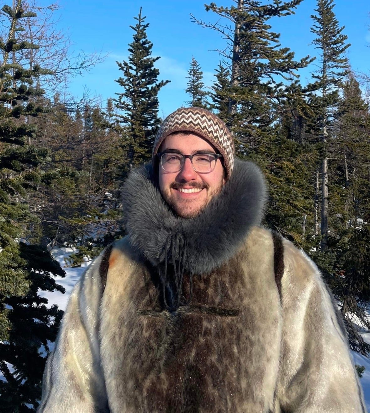 nicholas-flowers A man wearing a traditional Inuit winter coat smiles while standing in a forest.