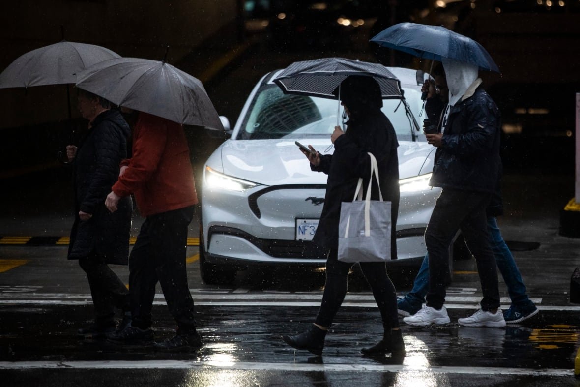 rain Four people with umbrellas walk in the rain on a street in front of a white car in the dark.