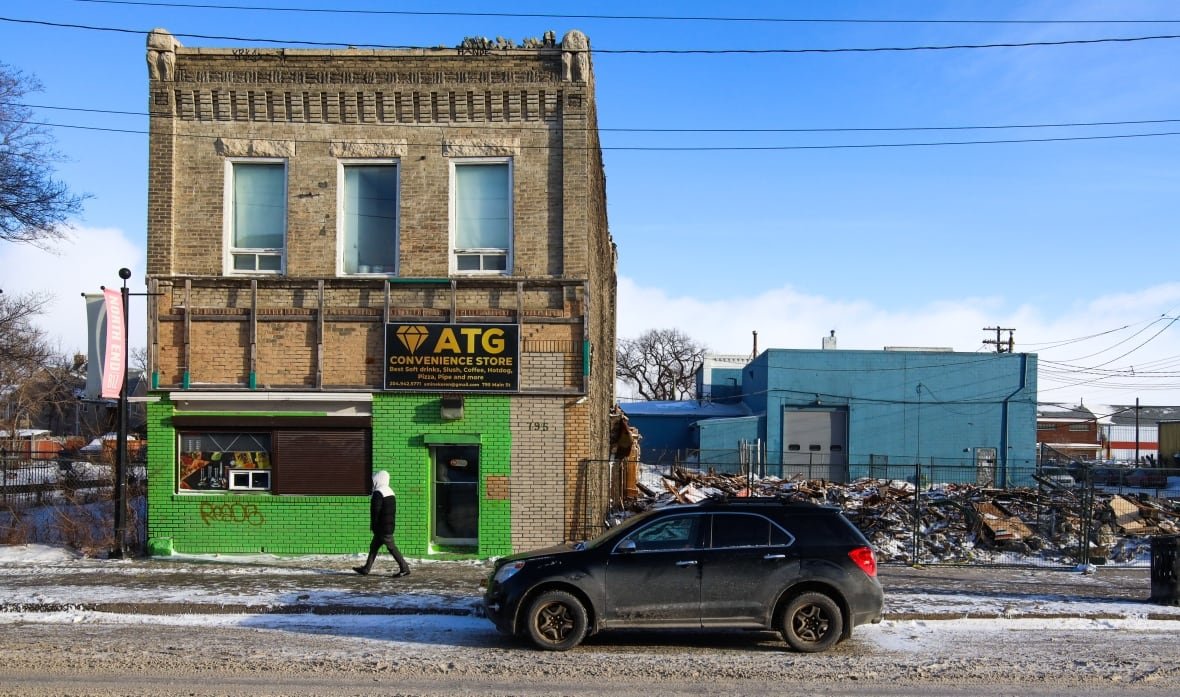 craig-building-alone-after-sutherland-hotel-burned A two-storey brick building with some decorative brick elements and its first storey painted bright green is on the left side of the photo. It sits between two lots without buildings. The one on the right is fenced off and full of rubble.