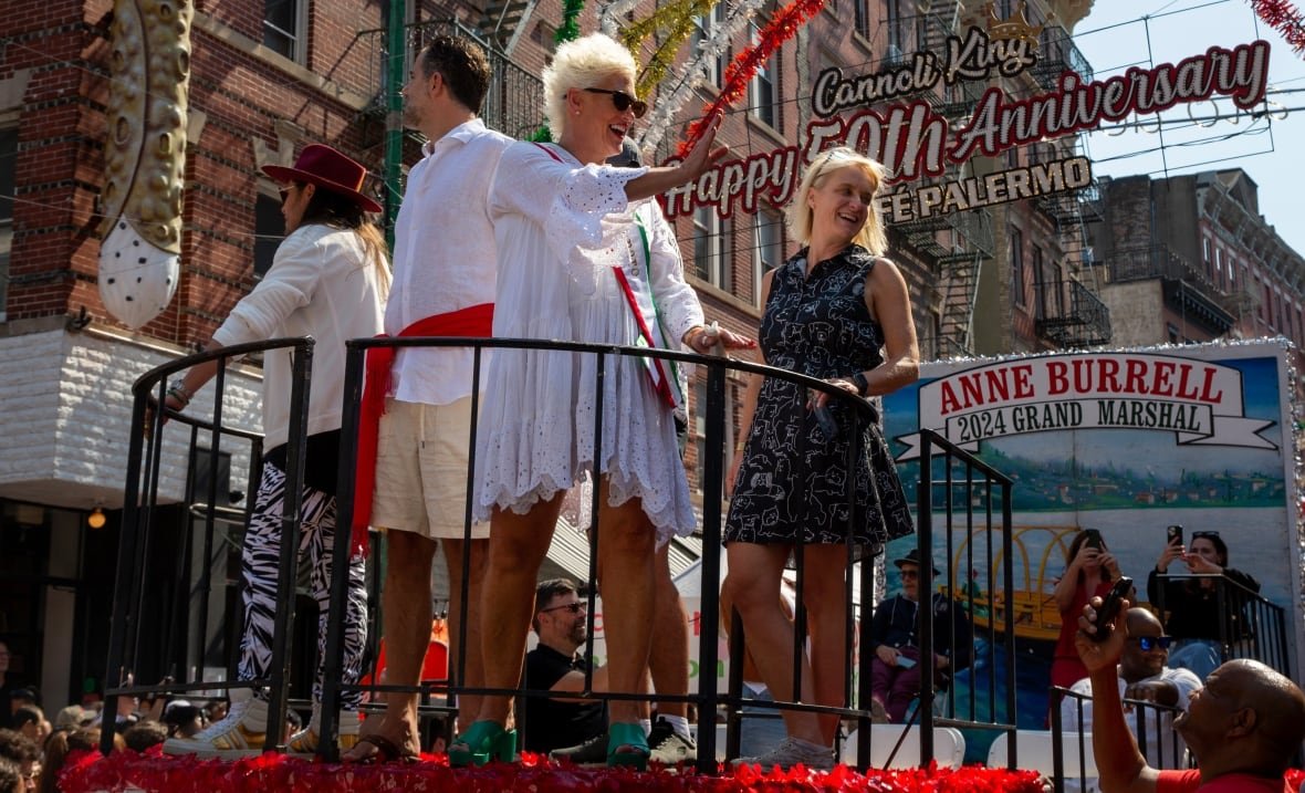anne-burrell A woman in a short white dress waves from the top of a parade float. There are four other people gathered on top of the float in a section hemmed in by a round railing.