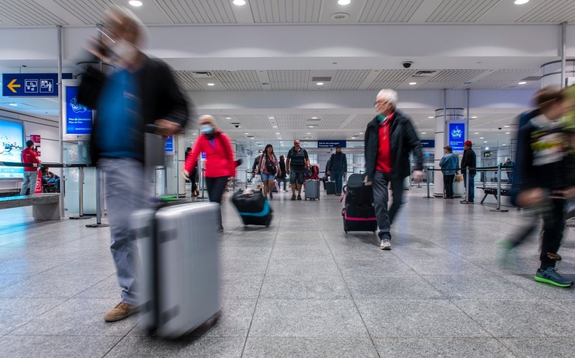 Blurred figures walk towards the camera in an airport terminal, wheeling luggage by their side.