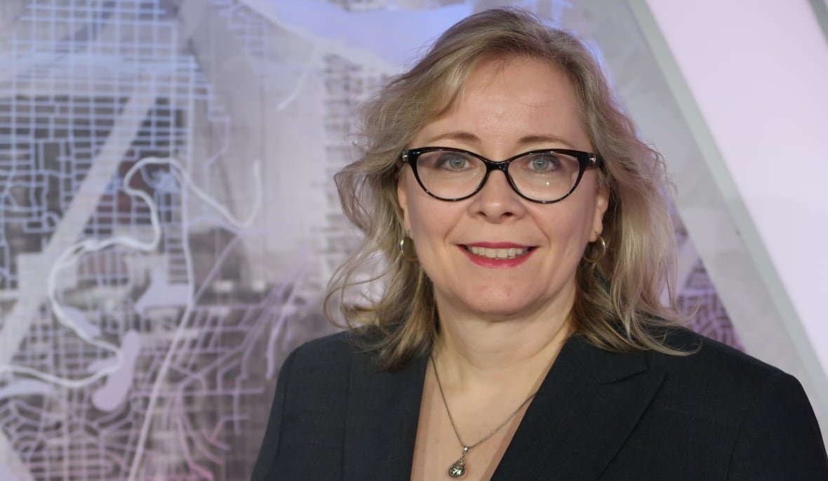 janet-brown A woman with blonde hair and glasses is smiling in front of a TV set, which shows a map of Calgary.