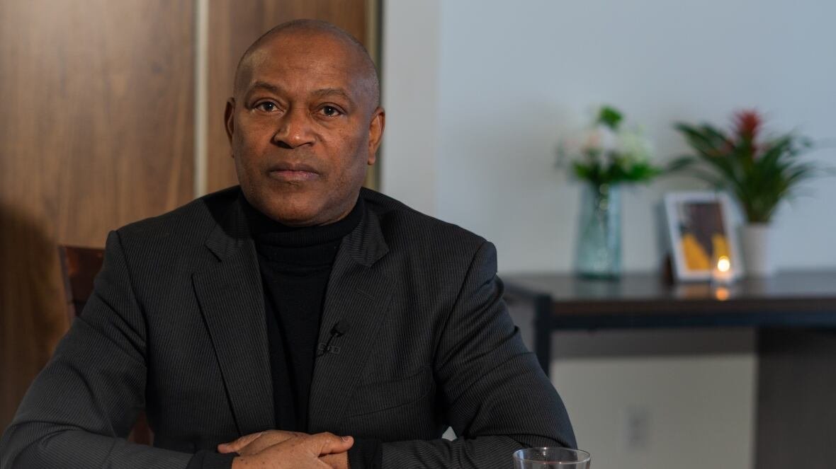 jean-dedieu-hakizimana A Black man in a blazer sits at a table with his arms folded.