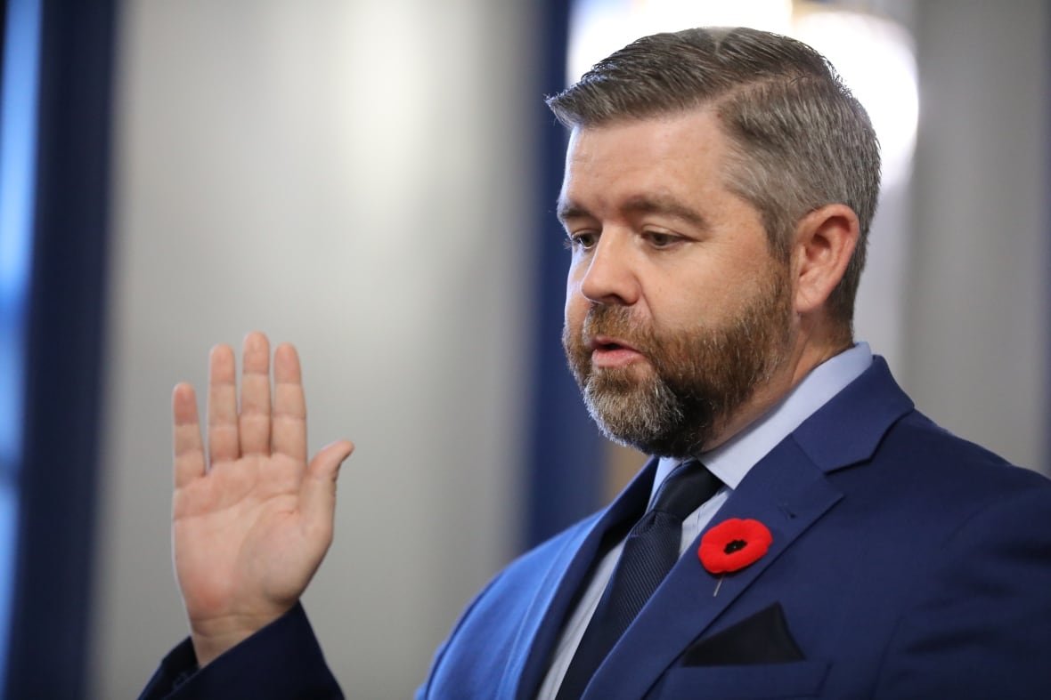 A bearded man raises his right hand. He is wearing a blue suit, white shirt and dark blue tie.