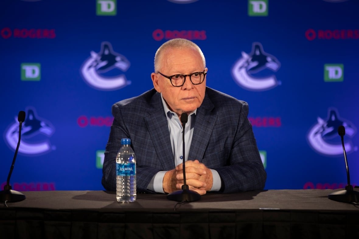 jim-rutherford-canucks A man in a dark suit and white open-collared shirt sits at a desk in front of a microphone with a plastic water bottle beside him, waiting for questions. Behind him is the Vancouver Canucks large-C-and-orca logo on a blue background.