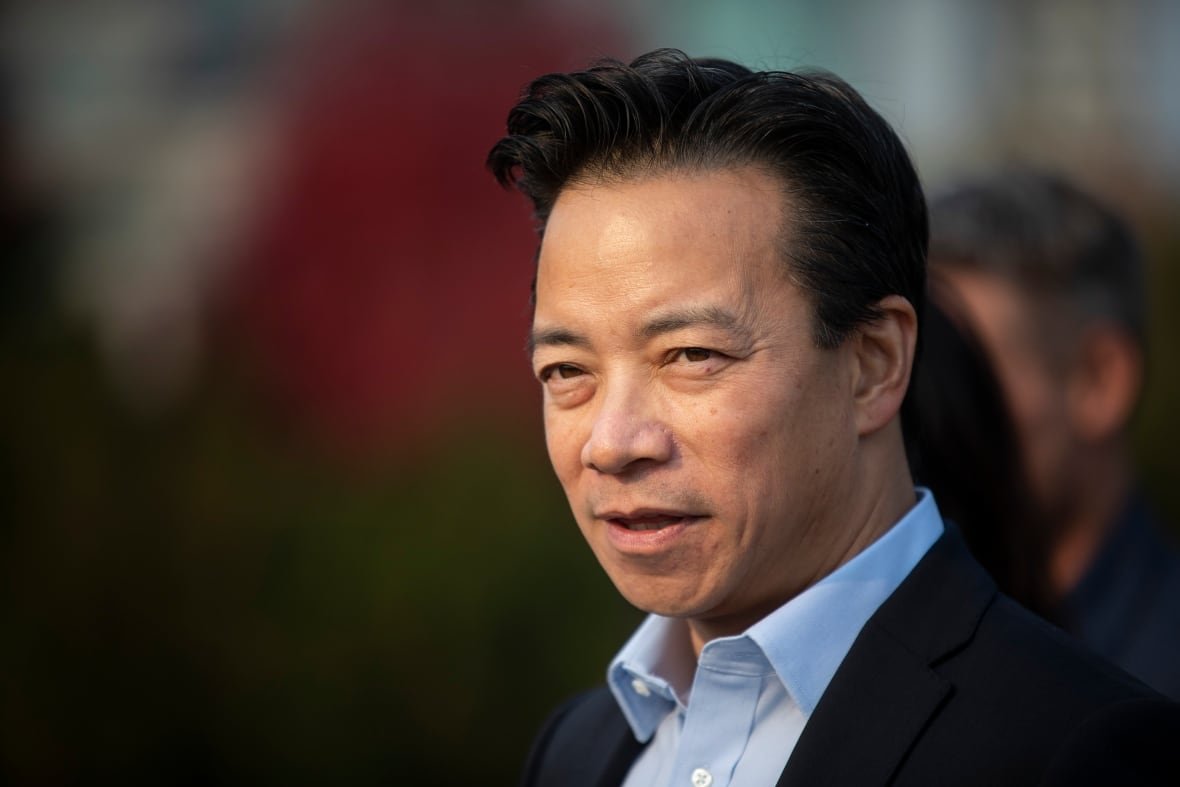 ken-sim-2 A close-up of a man in a dark suit and light blue shirt against a dark, blurred-out background.