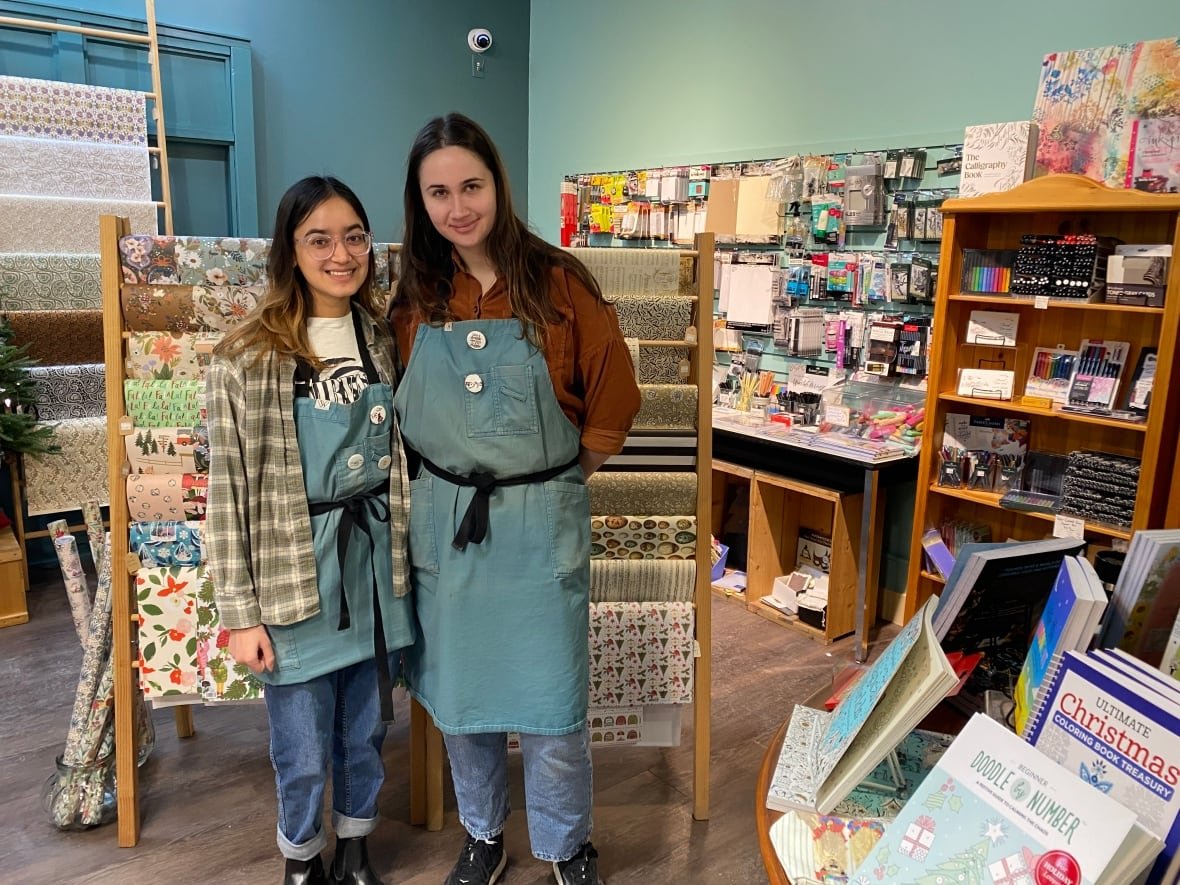 namya-and-mia Two women in blue smocks and jeans stand in the middle of a shop surrounded by paper andbooks.