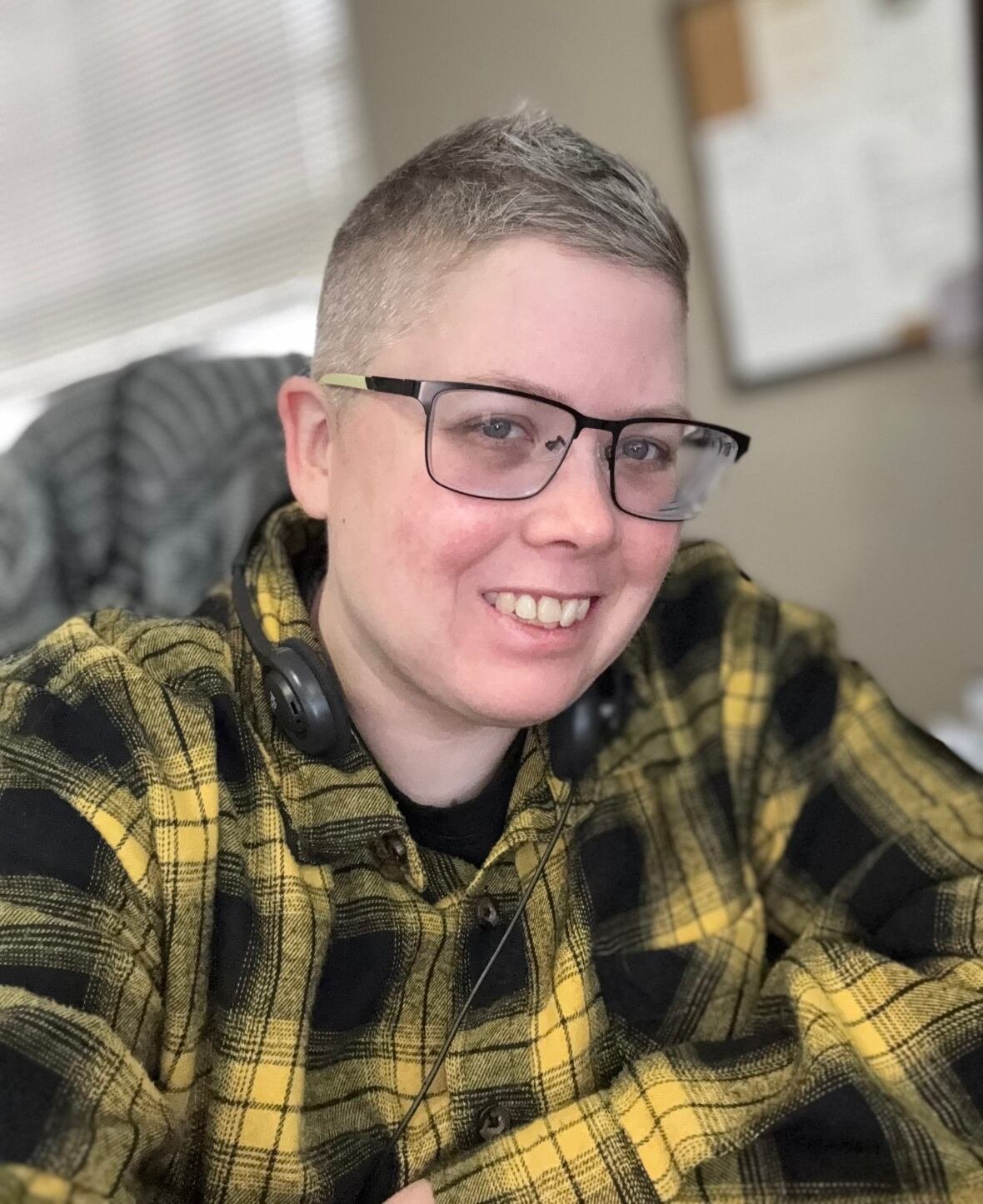 nicki-lyons-macfarlane A person with short, grey hair and wearing a yellow plaid jacket and glasses smiles.