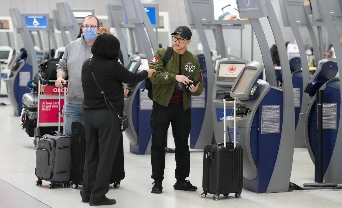 spring-break-travel-20230310 Two people stand near the kiosks at an airport, one pointing at the other, who is juggling a small object, maybe a passport. A third person is visible beyond them, wheeling a carry-on piece of luggage. Another piece of carry-on luggage sits near a kiosk.