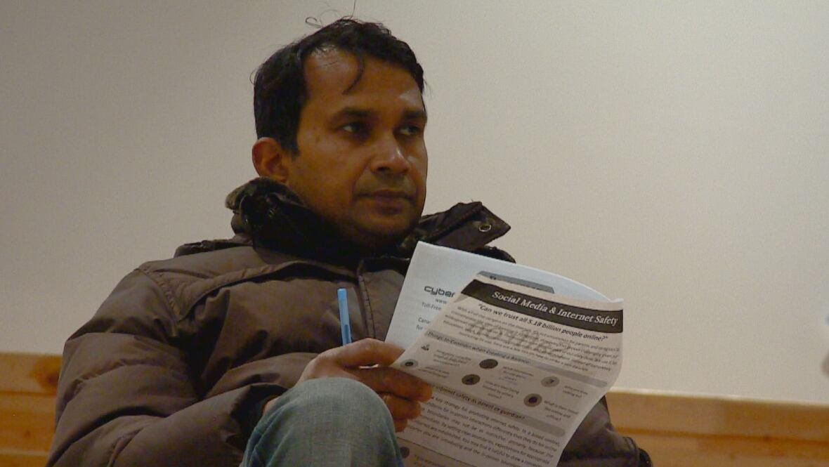 terrance-sambhudyal A man with short black hair and brown skin is sitting on a wooden bleacher. He is wearing a brown, puffy winter coat and blue jeans. He is holding paper booklet and a blue pen.