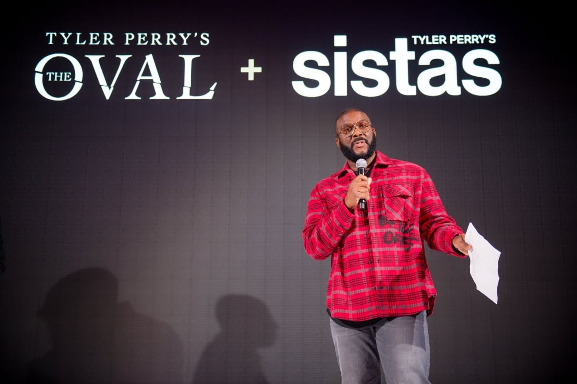 tyler-perry-the-oval A man in a red plaid shirt stands on a stage, holding a microphone in his right hand a and piece of paper in his left hand, in front of a black backdrop with the titles of two TV shows, The Oval and Sistas, printed in white letters.