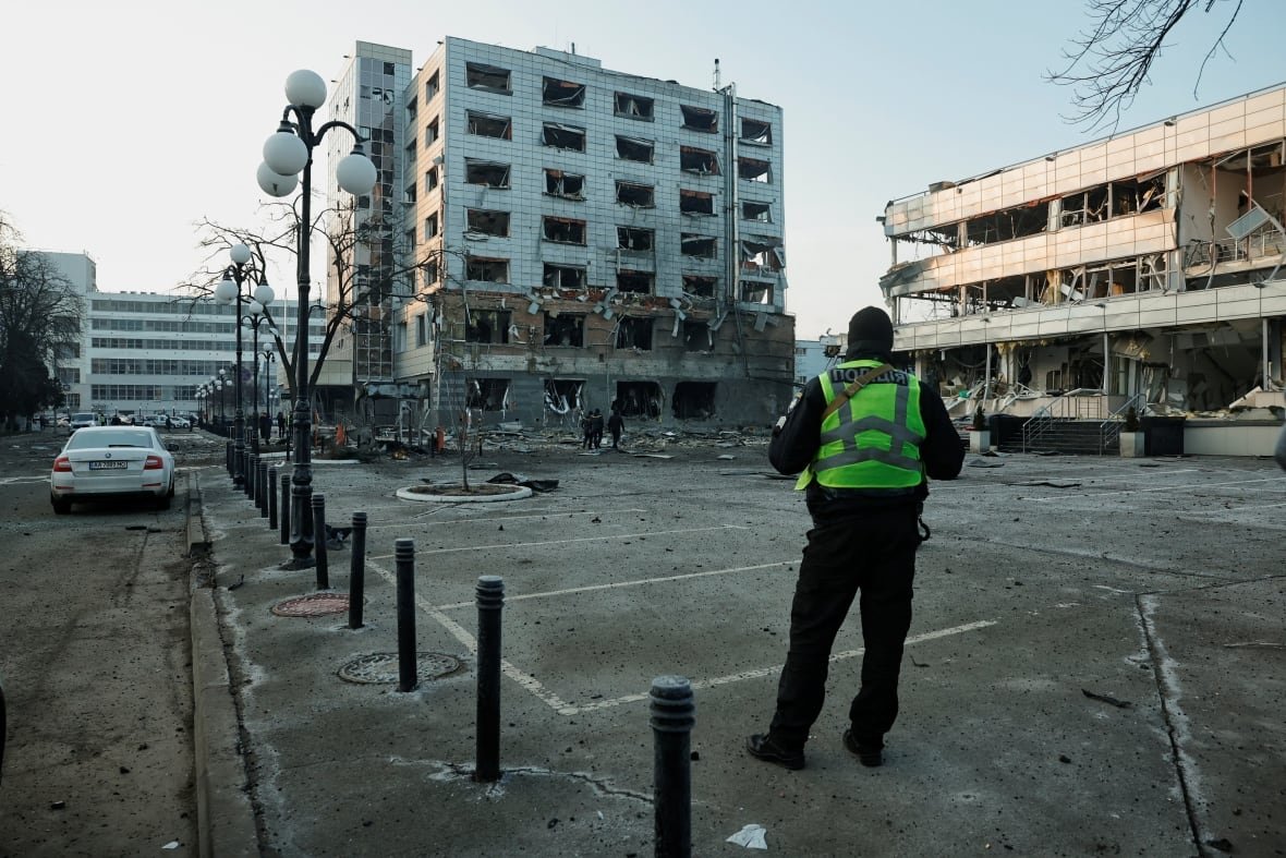 ukraine-crisis-attack-kyiv A police officer in a reflective vest with their back to the camera looks in the direction of two buildings, one a high-rise, heavily damaged.