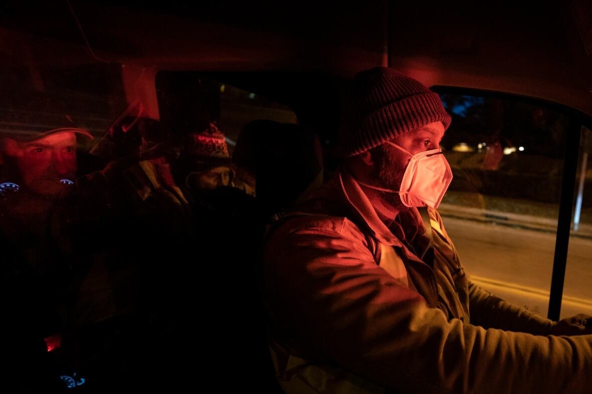 warming-shelter A man wears a medical mask and a toque while sitting in the driver's seat of a vehicle. It is dark outside and there is the silhouette of people sitting behind him in the vehicle.