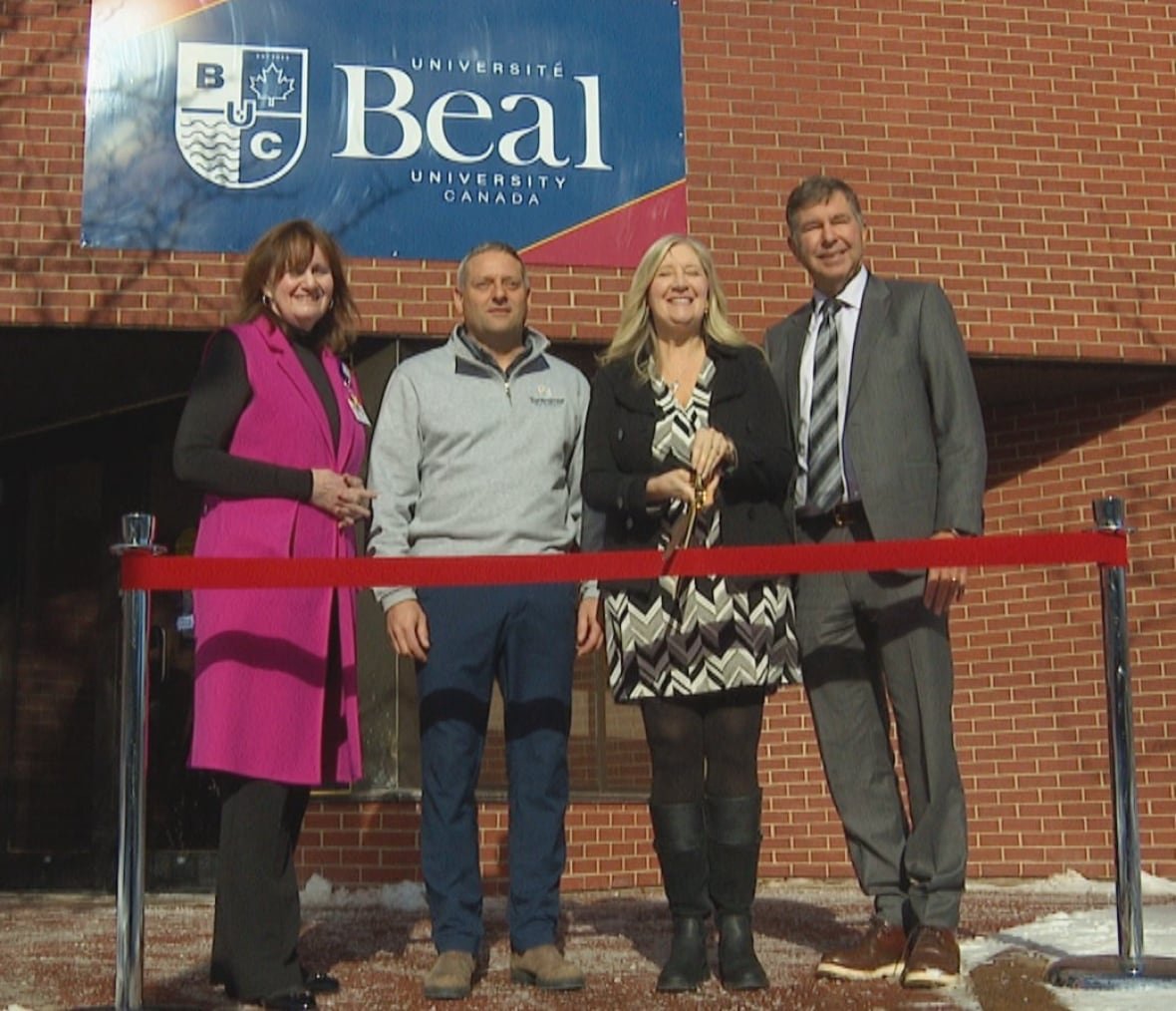 beal-university-sackville-opening A woman with short brown hair, a man wearing a grey sweater, a woman with long blond hair and a man in a grey suit stand behind a red ribbon. Behind them, a brick building has a sign that says "Beal"