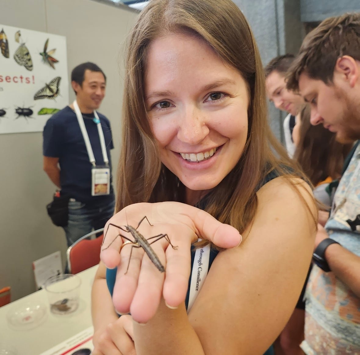katie-marshall A woman smiling at the camera holding a bug, with people looking at a bug exhibit in the background.