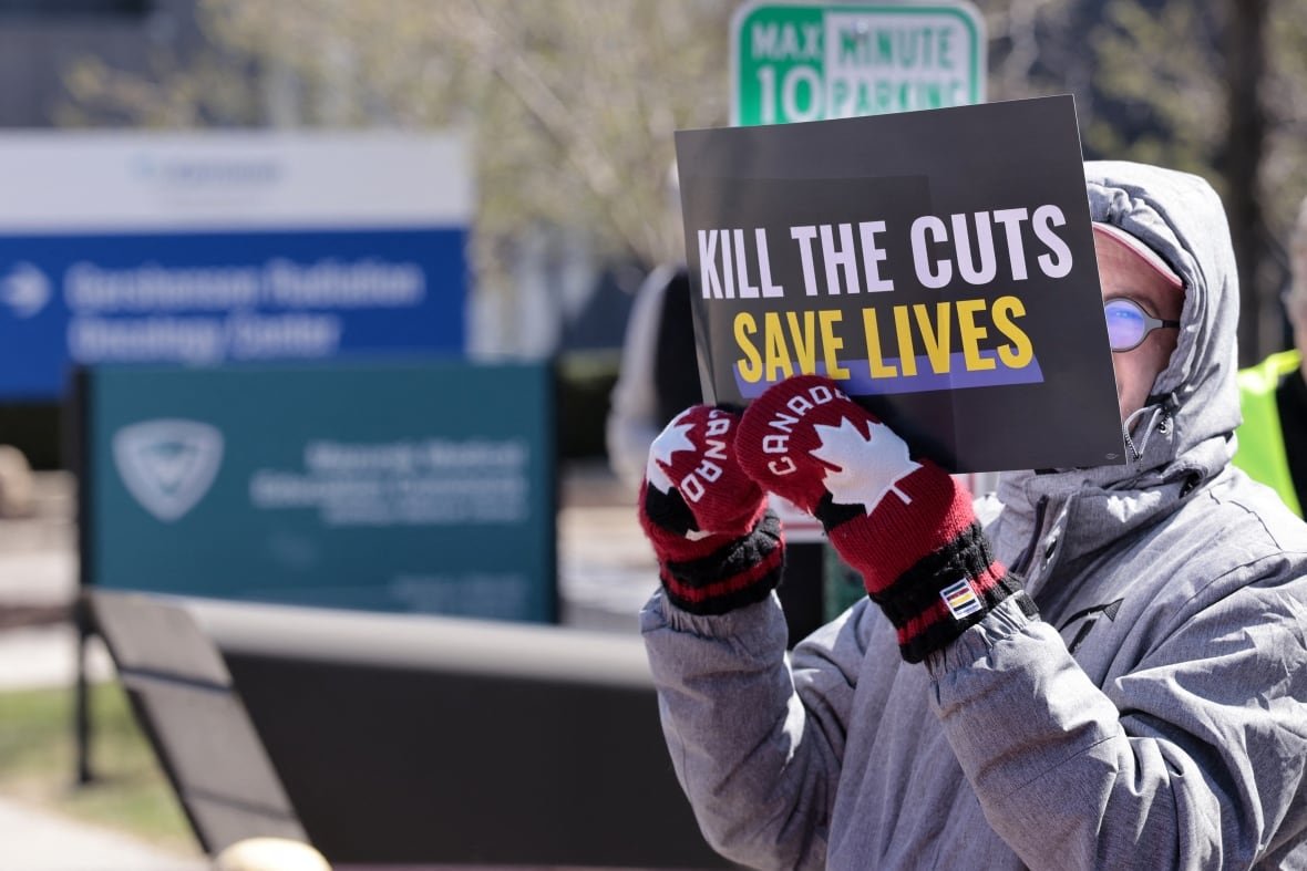 A person stands with a sign saying "Kill the Cuts" while wearing Canada mittens.