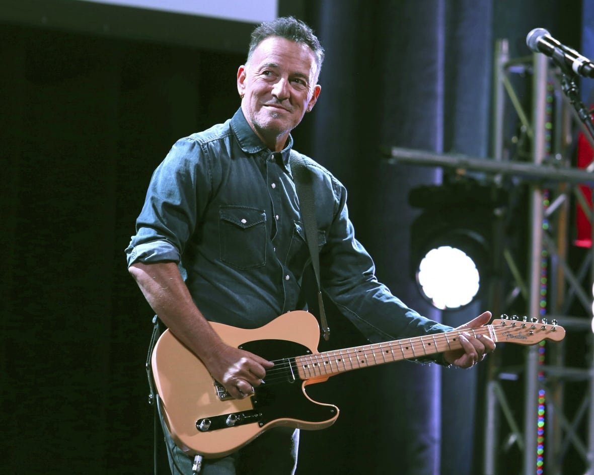 A man holding a guitar on stage looks to his right, smiling.
