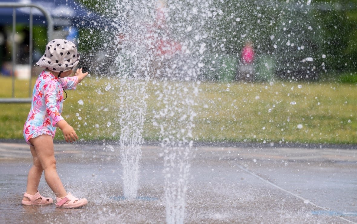 que-heat-wave-20250623 A toddler plays with fountain water at a park.