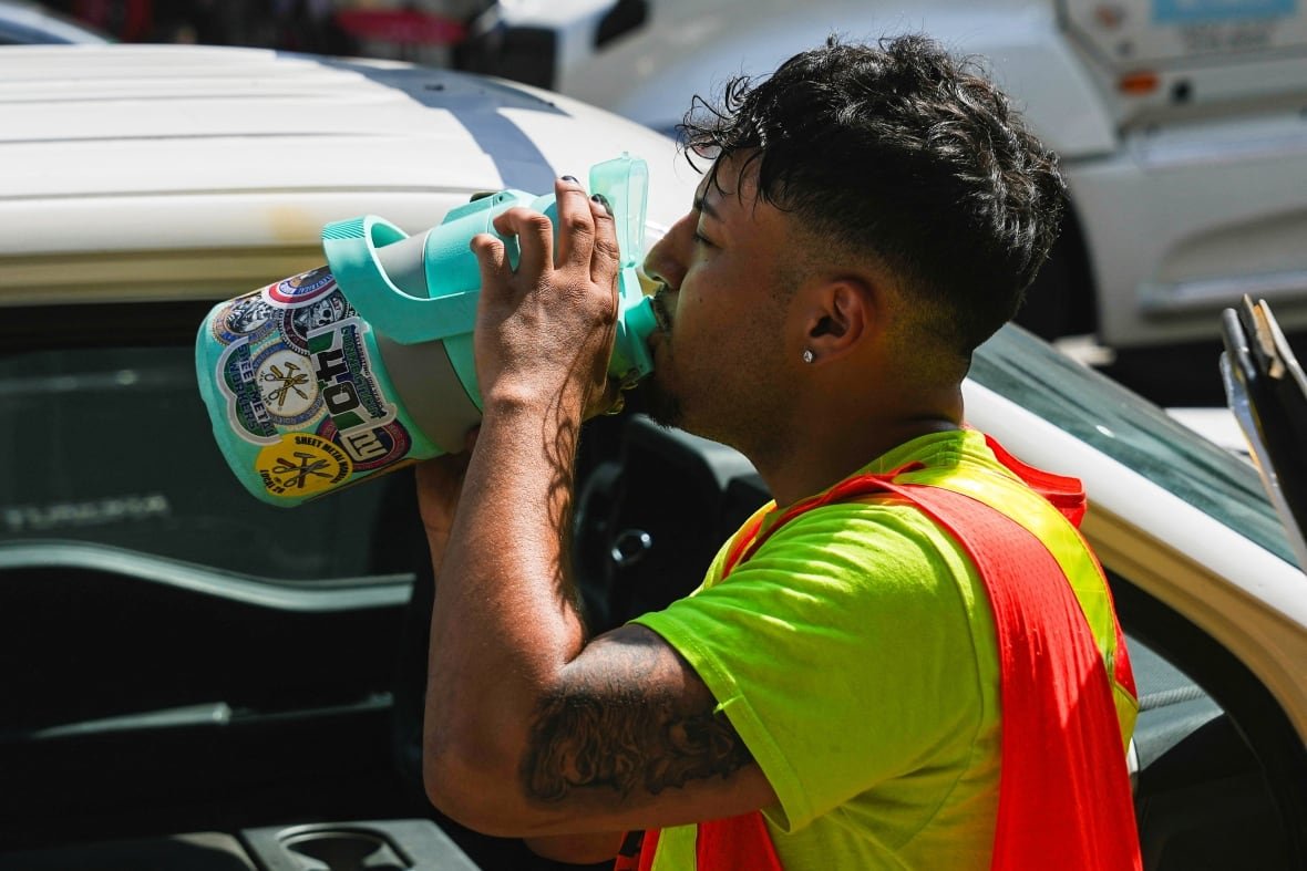 usa-weather-1 Wearing a yellow T-shirt and safety vest, a worker drinks from a giant canister near a vehicle, amid a heat wave.