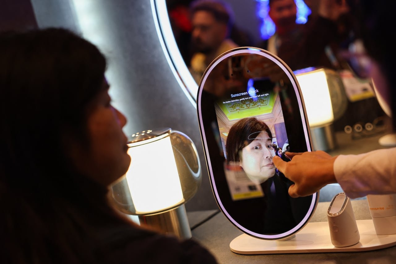 A person interacting with an oval screen resembling a mirror, with another individual's hand pointing at the reflection on the device.