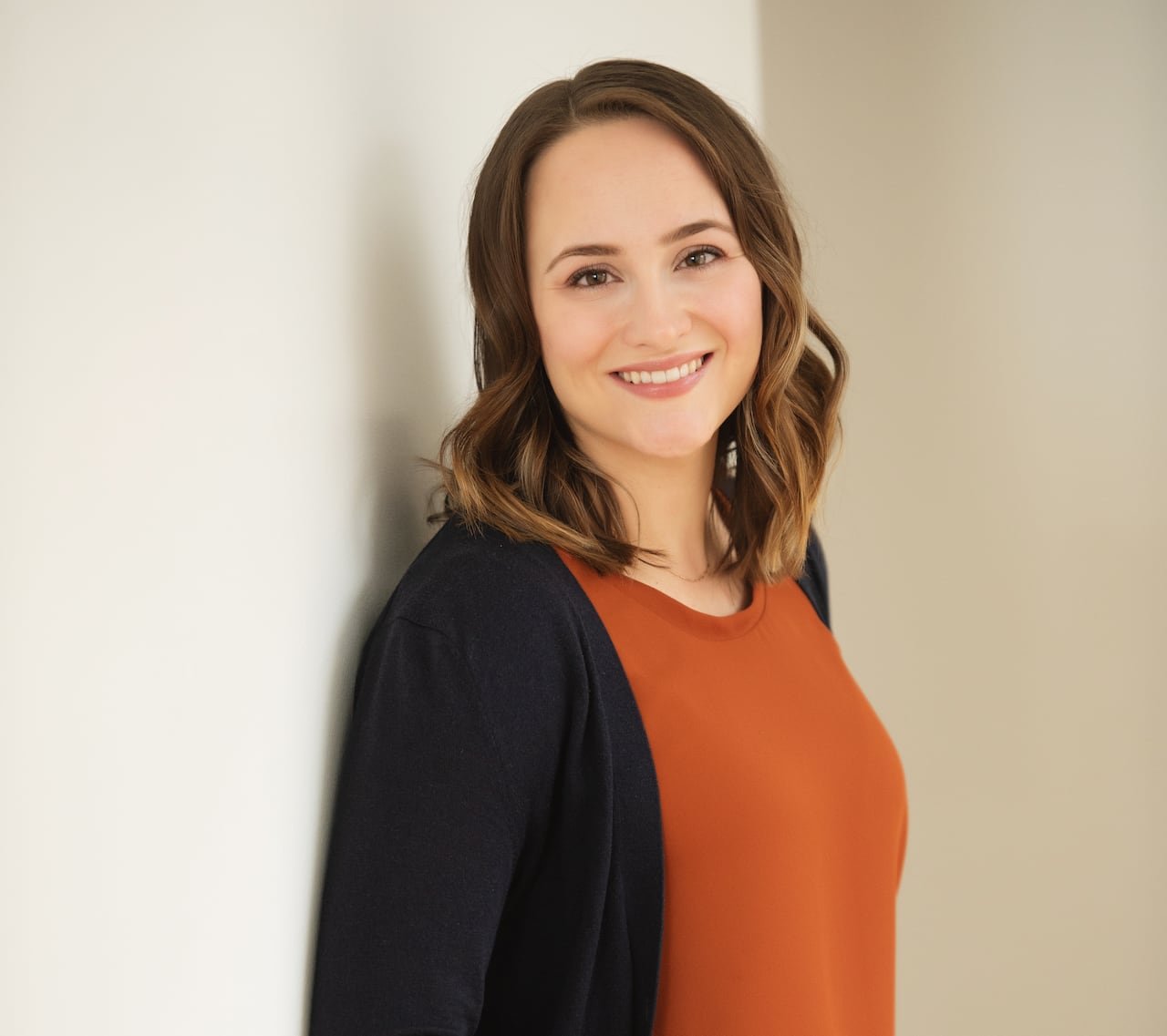 Woman wearing dark bottoms, orange top and dark cardigan stands smiling