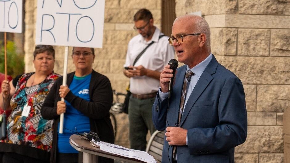 default-300 A politician speaks at an outdoor rally in autumn, with signs behind him that read 'no RTO.'