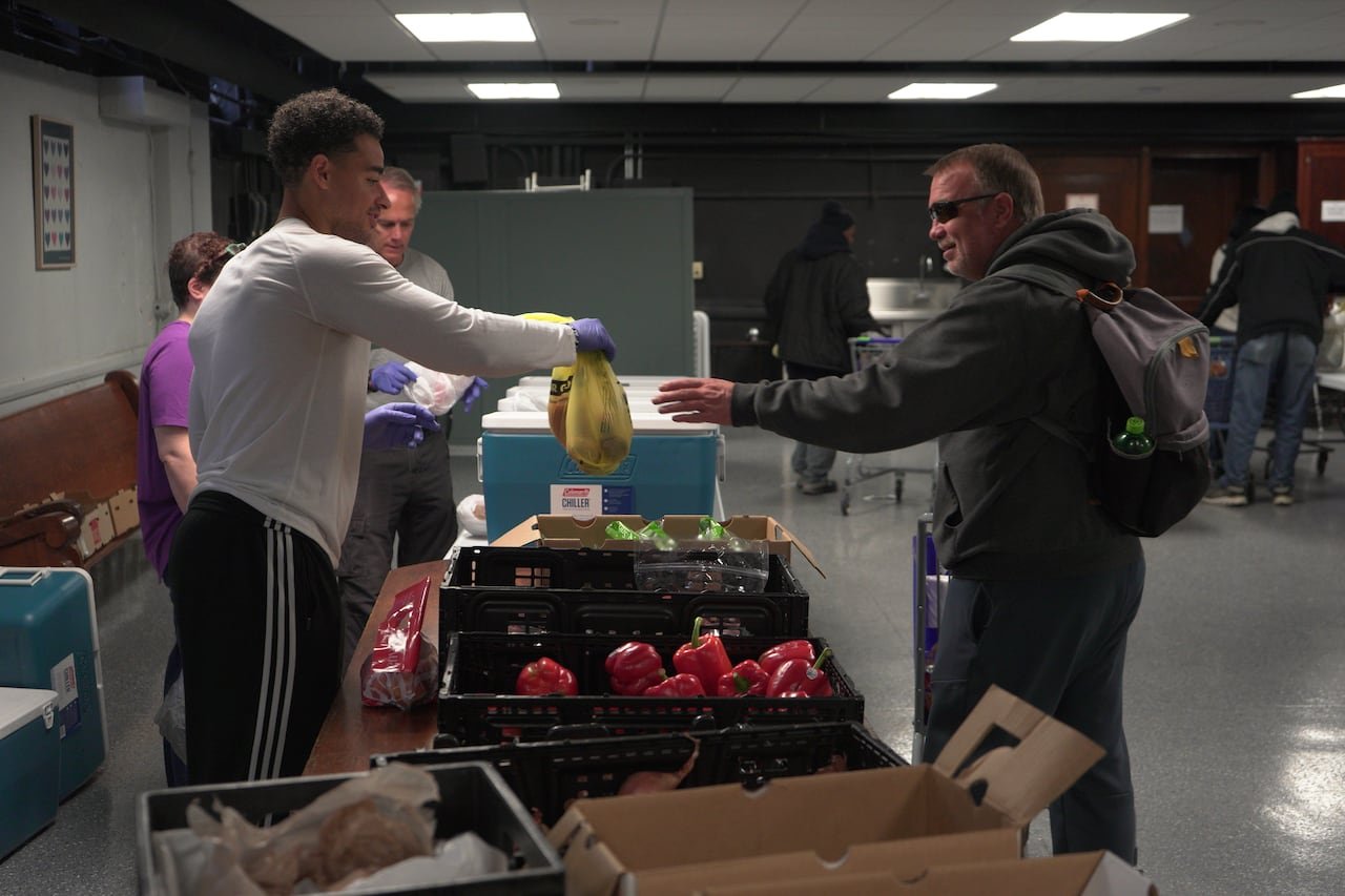 A volunteer handing a grocery bag to a man at a food bank, separated by a table with fruits and vegetables.
