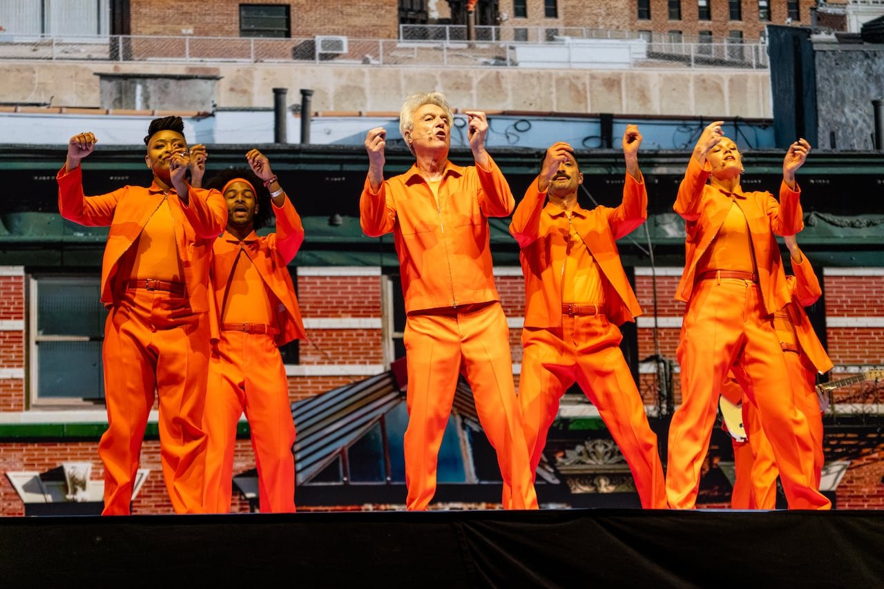 A male artist in orange attire performing with backup singers dressed similarly on stage.