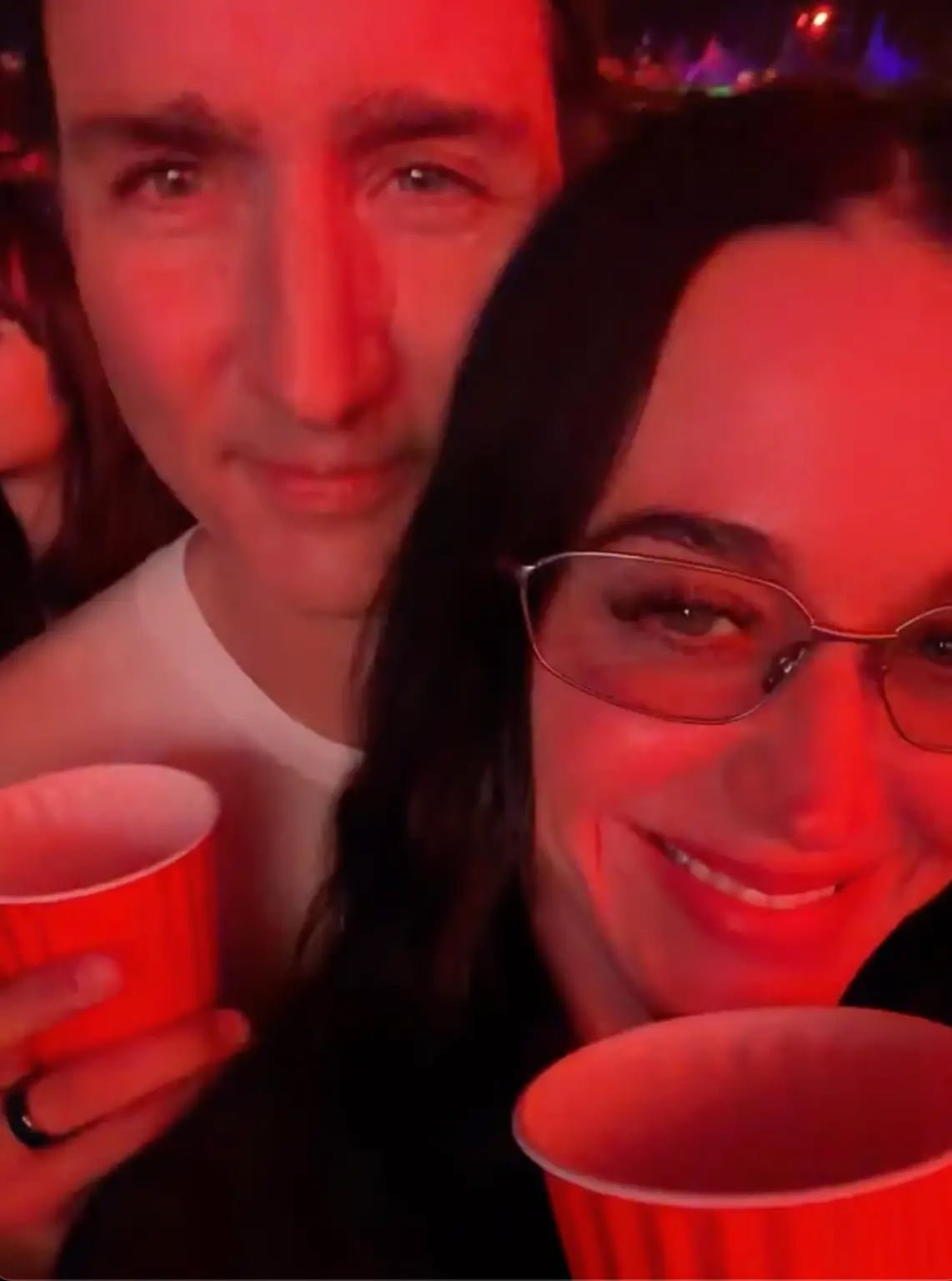 A man and woman taking a selfie at a music event, holding beverage cups and smiling.