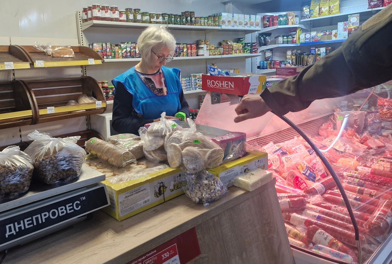 A woman wearing a blue smock stands behind a grocery counter holding bread, beans, and meat.