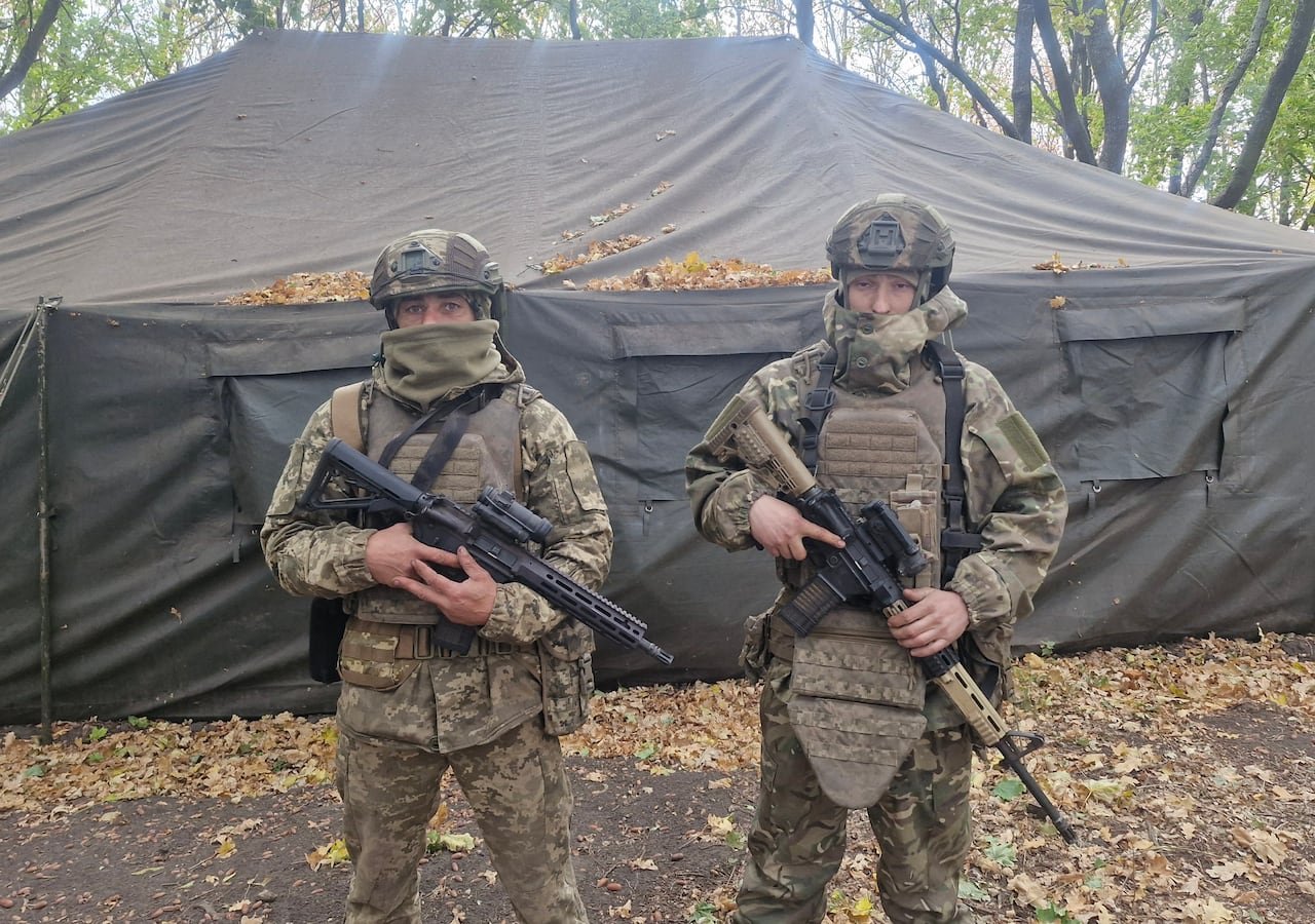 Two men in military fatigues stand in front of a large tent in a treed area, each holding a rifle.