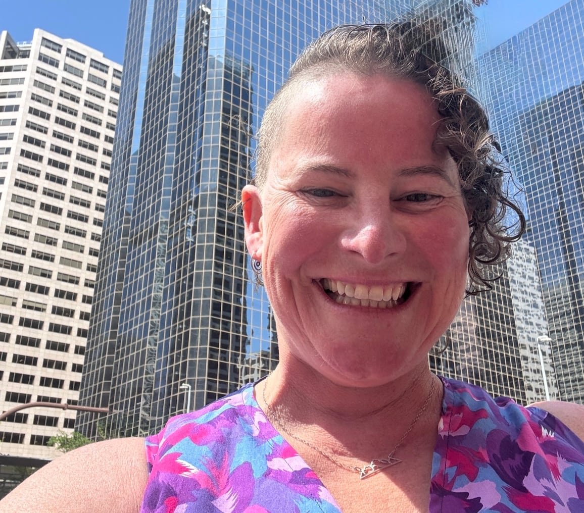 default-450 A woman with short curly hair shaved on one side smiles broadly with tall office buildings in the background.