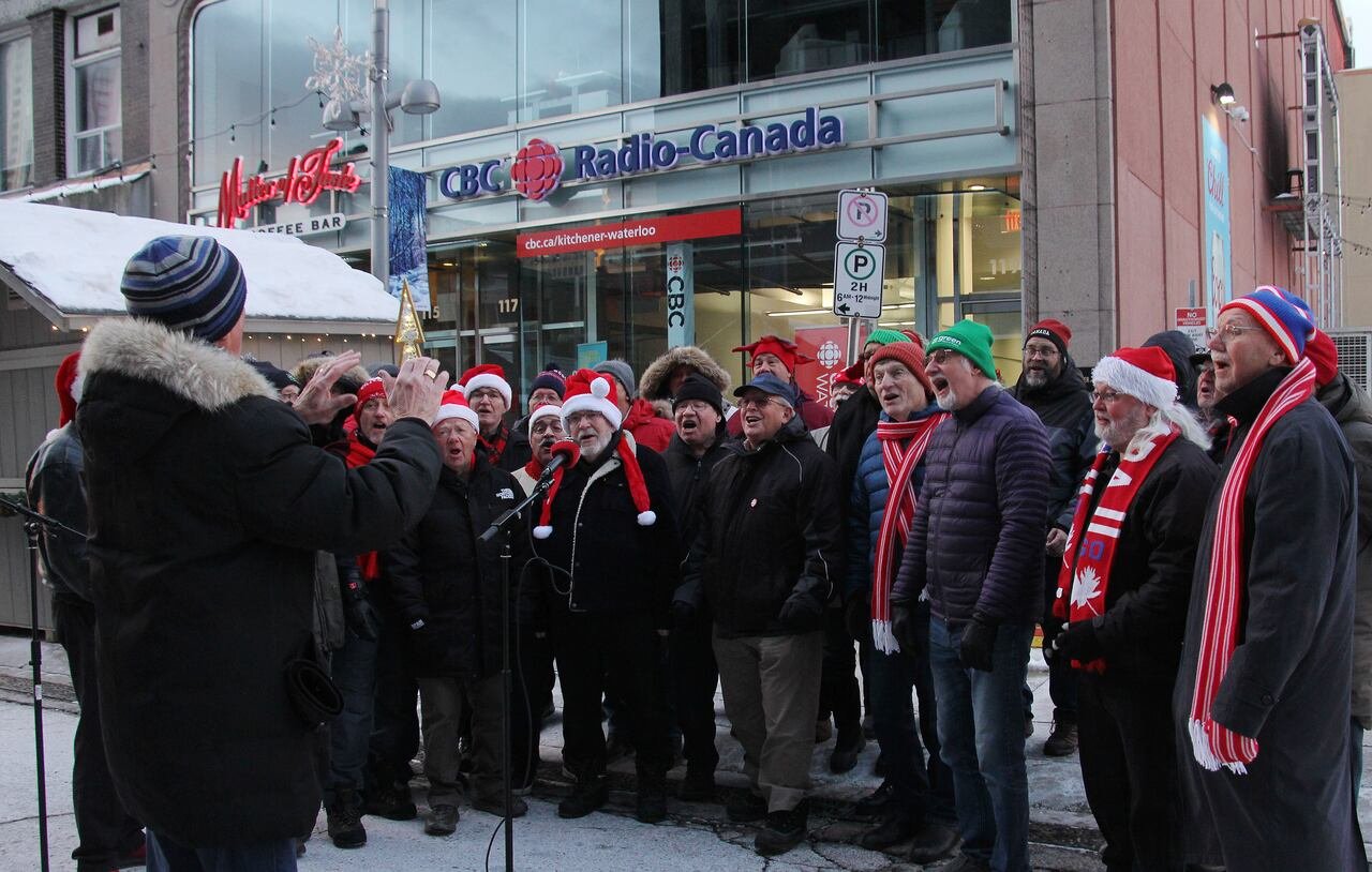 A group of about 24 men sing outside in cold weather in front of a CBC studio