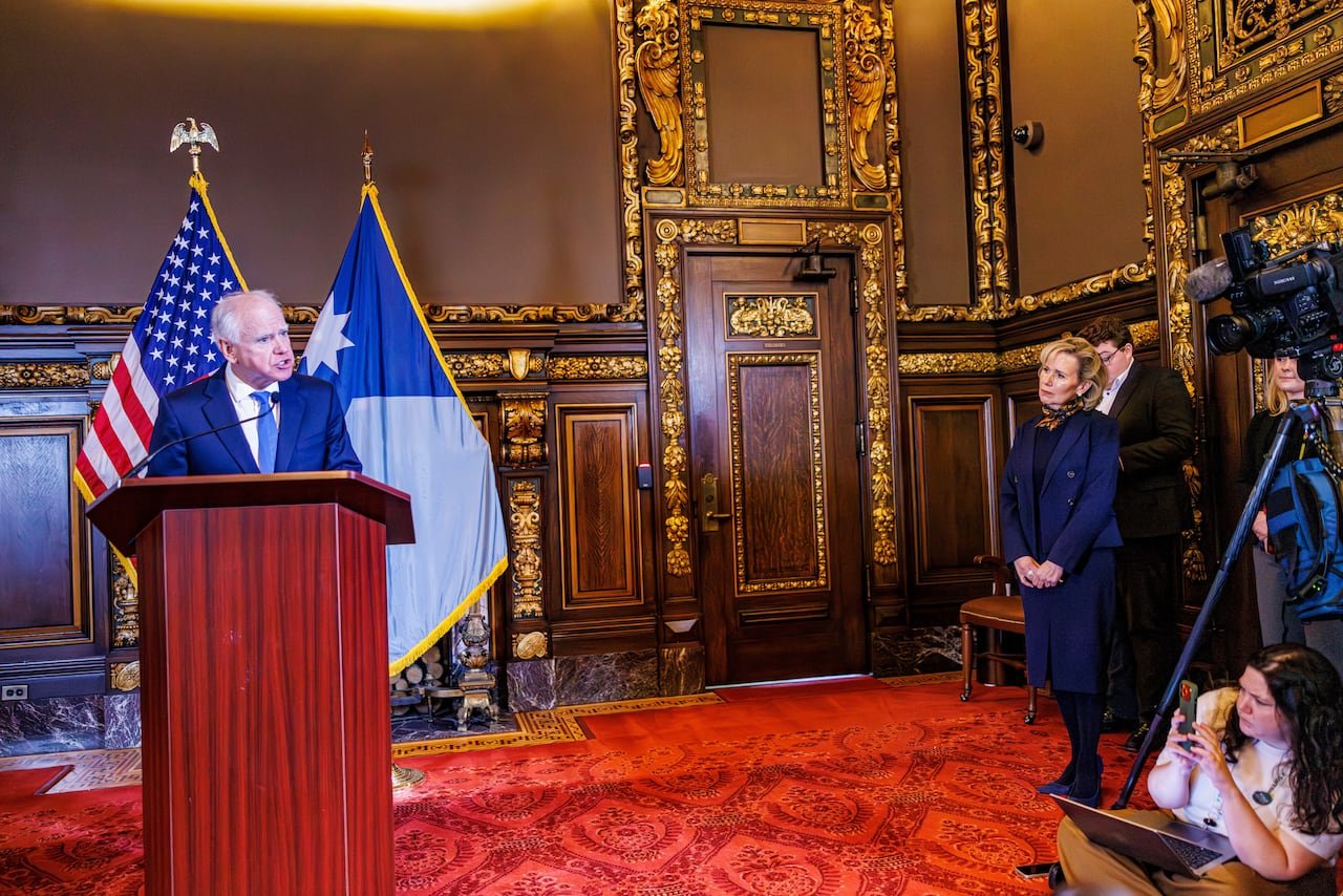 A person delivers a speech at a podium with others in the background.