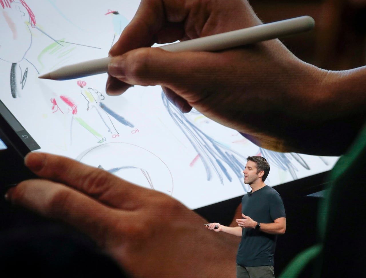 a man in a plain black tshirt stands on a stage introducing a new piece of technology. on the screen behind him, a child draws a picture using a stylus on an ipad